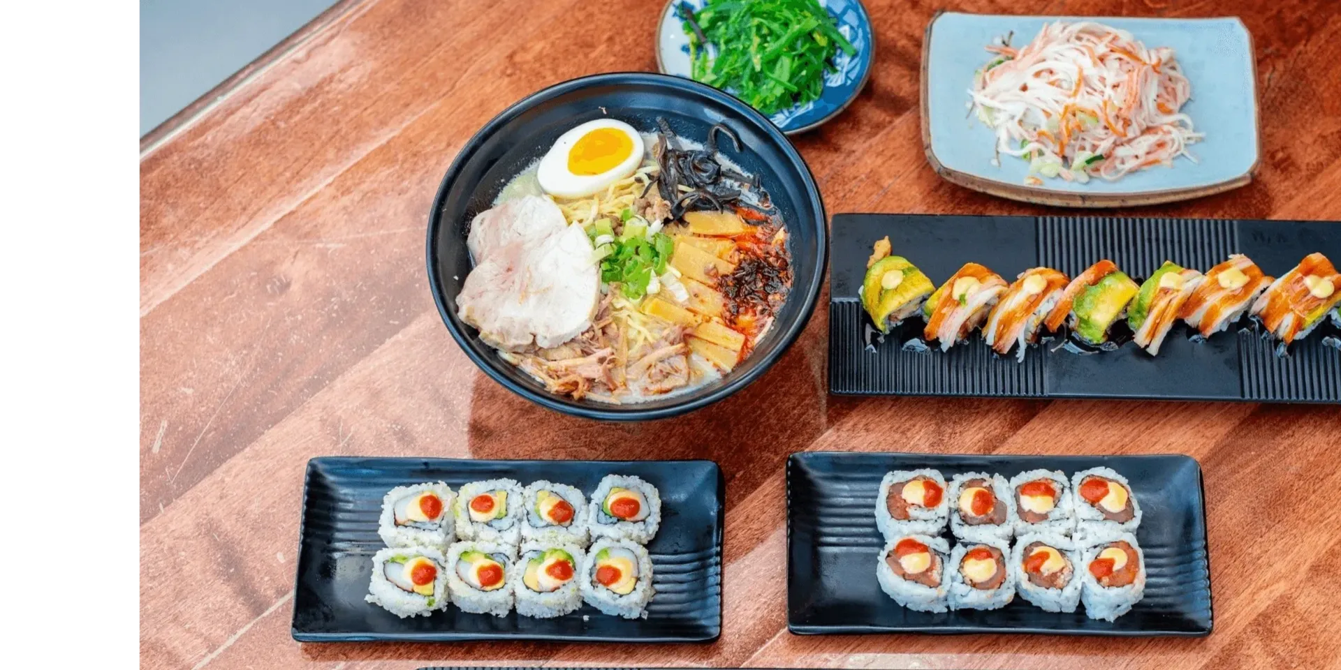 Japanese feast featuring ramen, assorted sushi rolls, seaweed salad, and kani salad on a wooden table.